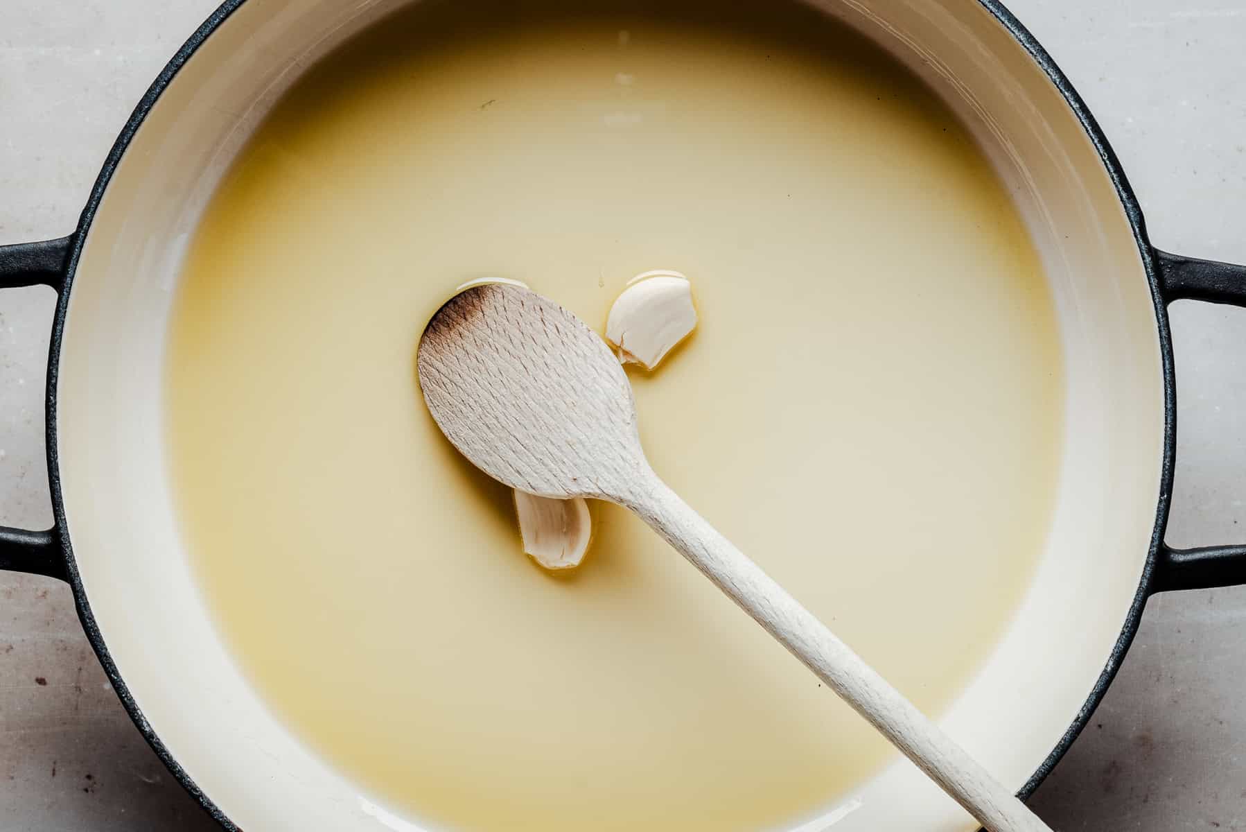 A wooden spoon rests in a pot filled with light yellow oil and two garlic cloves, viewed from above.
