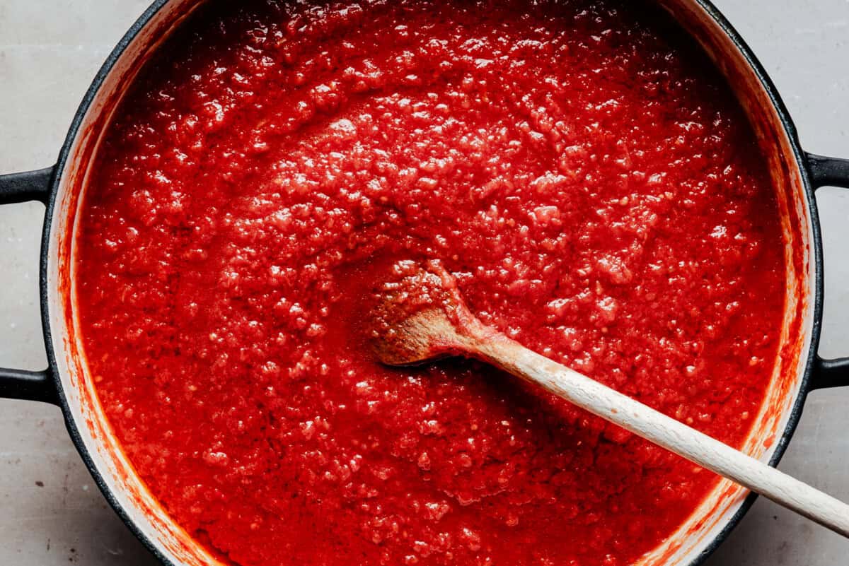 A wooden spoon stirs a pot filled with thick, vibrant red pomodoro sauce.