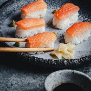 A plate of salmon nigiri sushi with wasabi and pickled ginger, with one piece being picked up by chopsticks. A small bowl of soy sauce is visible in the foreground.