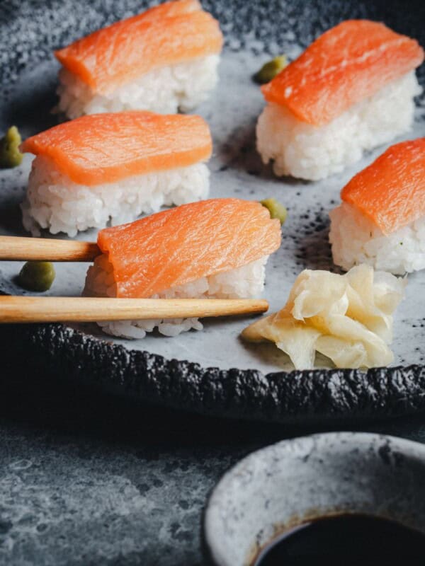 A plate of salmon nigiri sushi with wasabi and pickled ginger, with one piece being picked up by chopsticks. A small bowl of soy sauce is visible in the foreground.