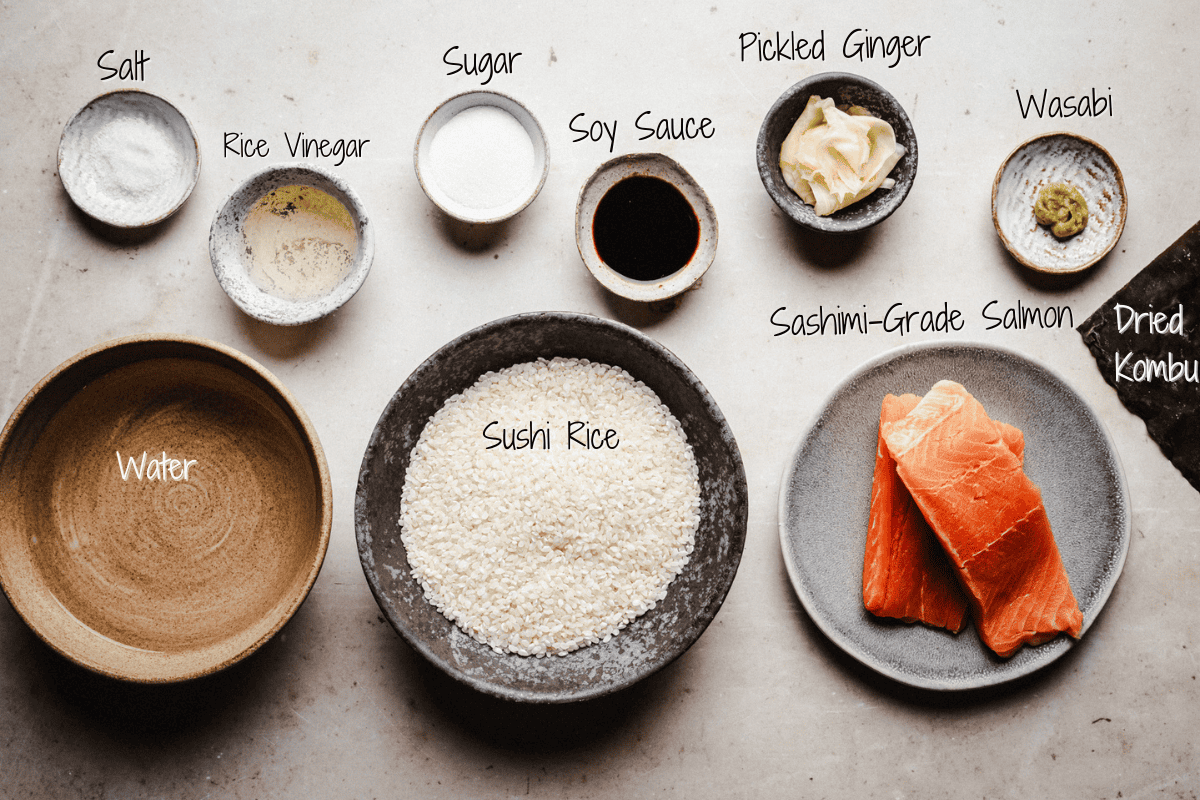 A flat lay of ingredients for salmon nigiri: salt, rice vinegar, sugar, soy sauce, pickled ginger, wasabi, dried kombu, water, sushi rice, and sashimi-grade salmon, each labeled in handwritten text.