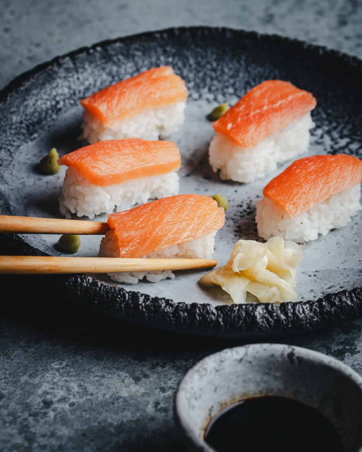 A black plate with five pieces of salmon nigiri sushi, small portions of wasabi, pickled ginger, and a pair of wooden chopsticks. A small dish of soy sauce is nearby on a gray surface.
