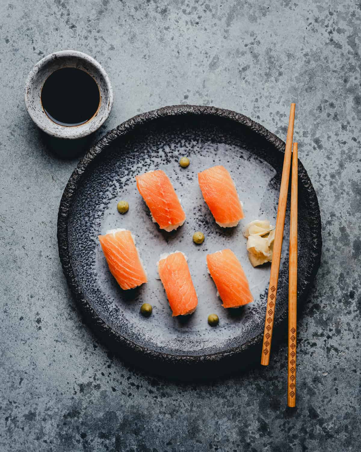 Six pieces of salmon nigiri sushi are arranged on a black plate with wasabi dots and pickled ginger, accompanied by chopsticks and a small bowl of soy sauce on a gray textured surface.