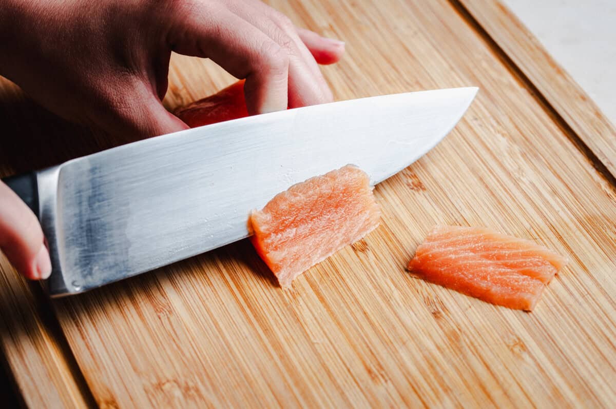 A close-up of a hand slicing raw salmon on a wooden cutting board with a large kitchen knife.