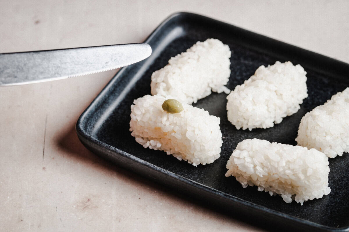 Five pieces of plain sushi rice are arranged on a black rectangular plate, with a small dab of wasabi on one piece. A serrated knife is visible near the plate's edge.