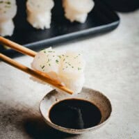 A pair of chopsticks holds a piece of scallop nigiri topped with white fish and chopped herbs over a small bowl of soy sauce, with more sushi on a tray in the background.