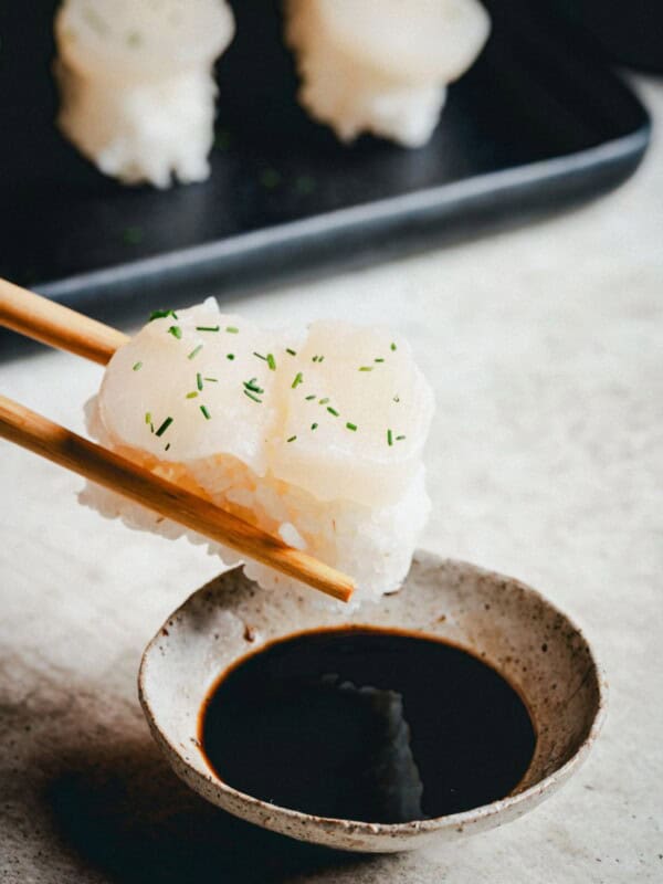 A pair of chopsticks holds a piece of scallop nigiri topped with white fish and chopped herbs over a small bowl of soy sauce, with more sushi on a tray in the background.