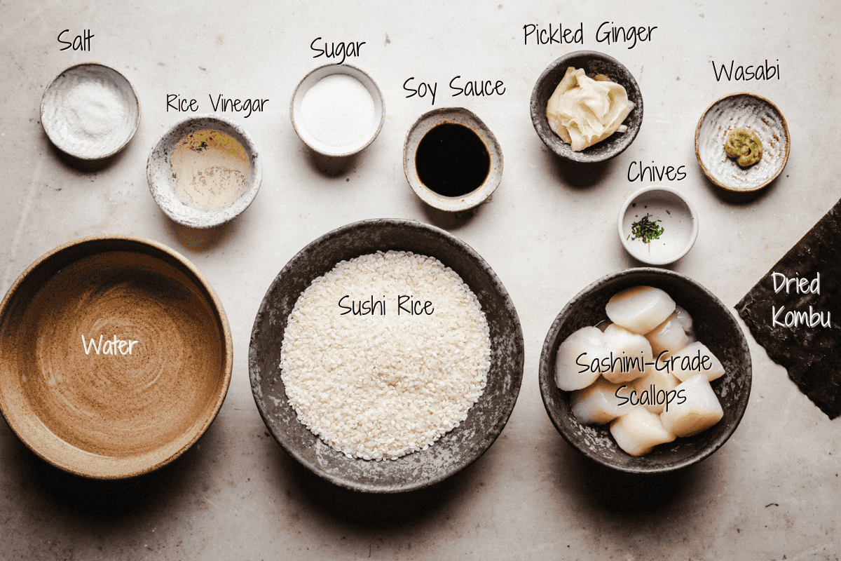 A top view of labeled bowls containing ingredients for scallop nigiri: salt, rice vinegar, sugar, soy sauce, pickled ginger, wasabi, chives, dried kombu, water, sushi rice, and sashimi-grade scallops.