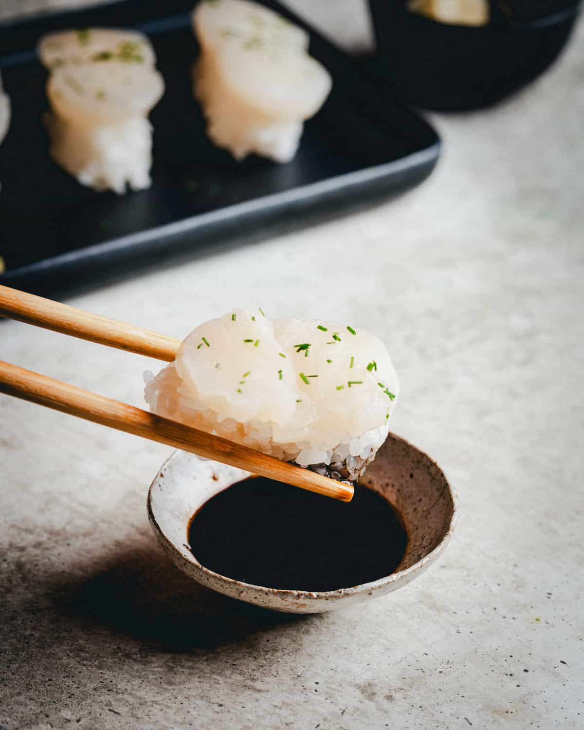 A pair of chopsticks holds a piece of scallop nigiri above a small bowl of soy sauce, with more nigiri pieces blurred in the background.