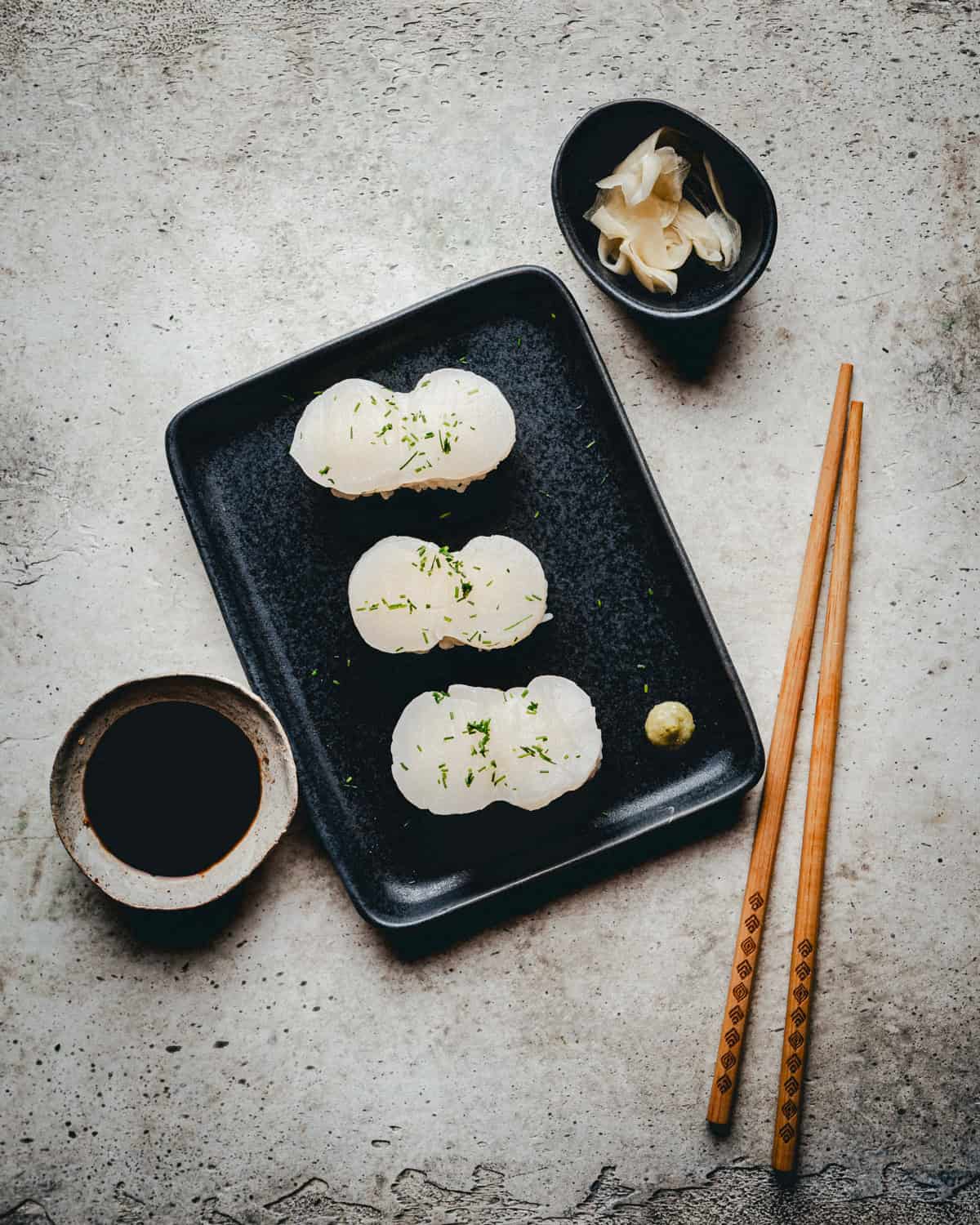 A black plate with six pieces of scallop nigiri garnished with green herbs, a small ball of wasabi, a bowl of soy sauce, pickled ginger, and a pair of wooden chopsticks on a light textured surface.
