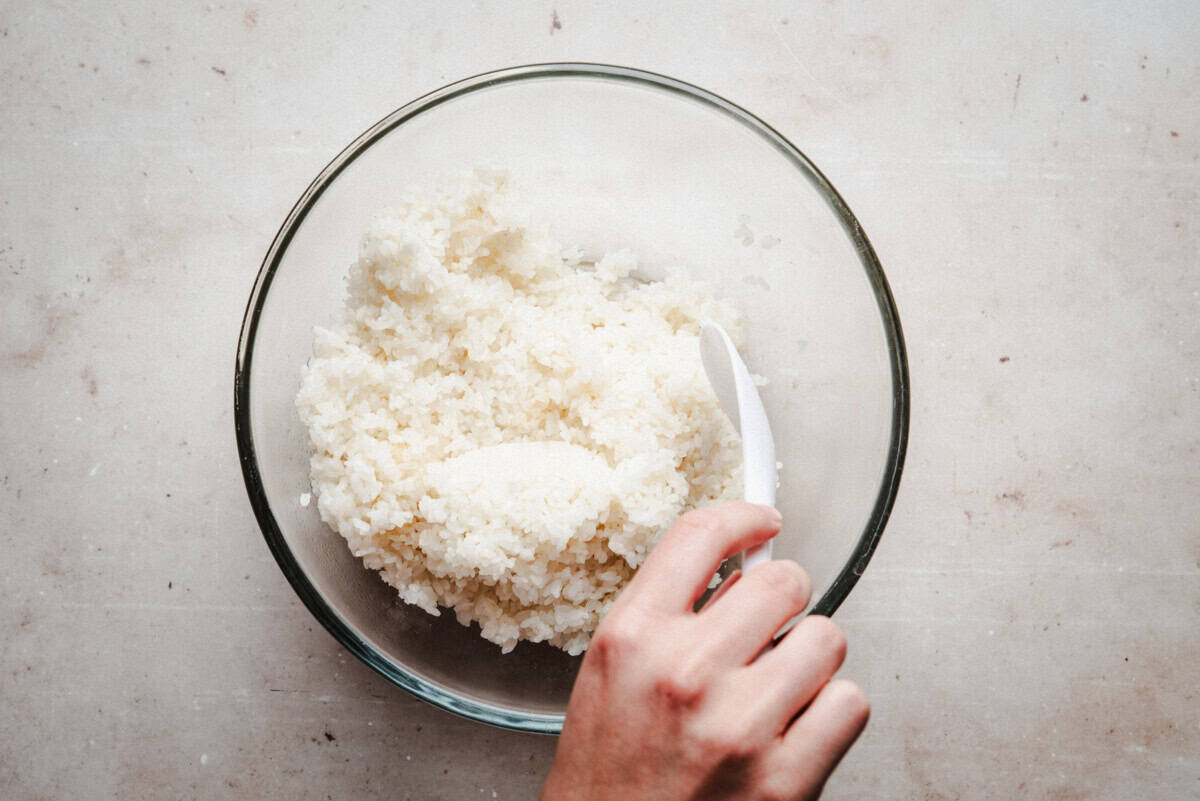A hand holding a white spoon, mixing or scooping white rice in a clear glass bowl on a light-colored surface.