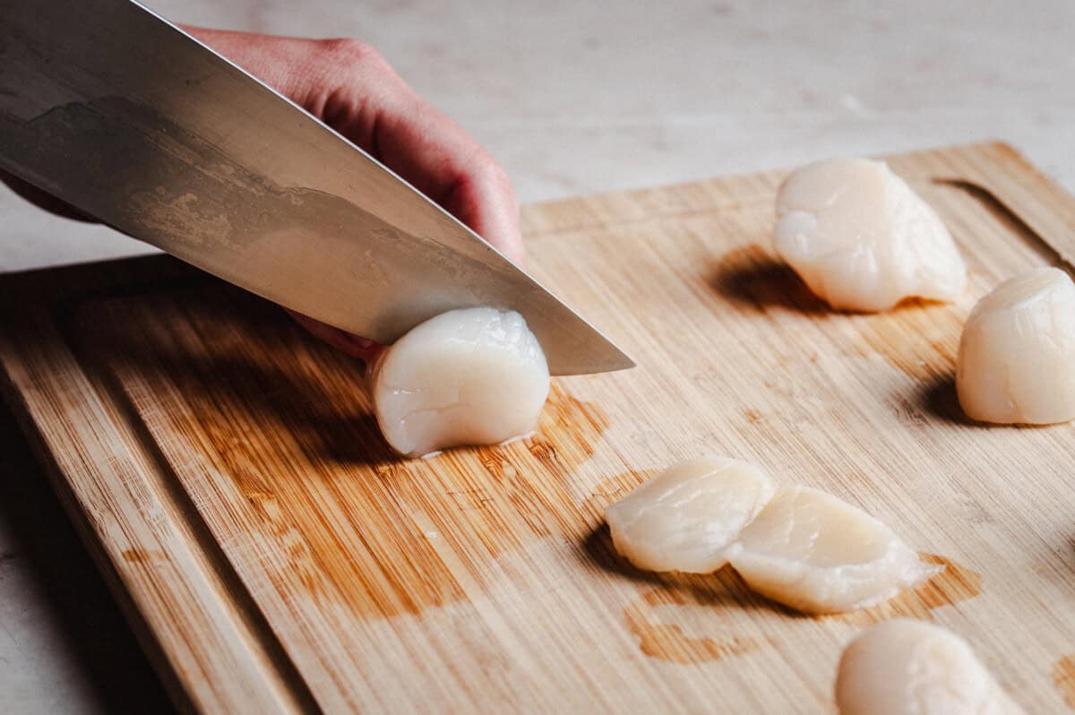 A hand holding a knife slices raw scallops on a wooden cutting board, with several whole and sliced scallops arranged nearby.