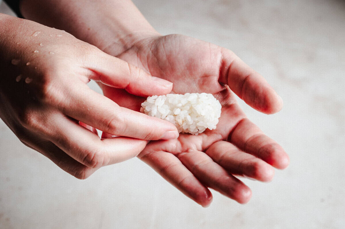 A close-up of two hands shaping a small mound of white rice, with a few grains of rice and water droplets visible on the fingers.