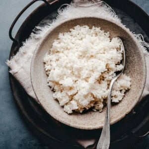 A rustic ceramic bowl filled with cooked sushi rice sits on a cloth napkin with a metal spoon, placed on top of a round tray.