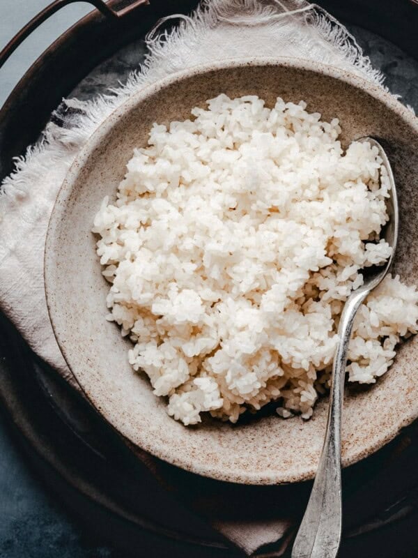 A rustic ceramic bowl filled with cooked sushi rice sits on a cloth napkin with a metal spoon, placed on top of a round tray.
