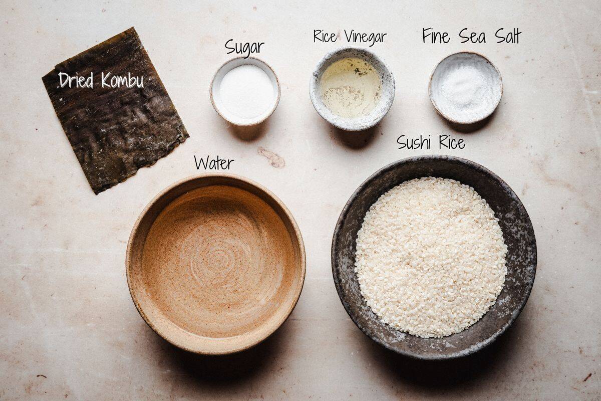 Bowls containing sugar, rice vinegar, fine sea salt, water, and sushi rice, along with a piece of dried kombu, are arranged on a light surface; each ingredient is labeled.