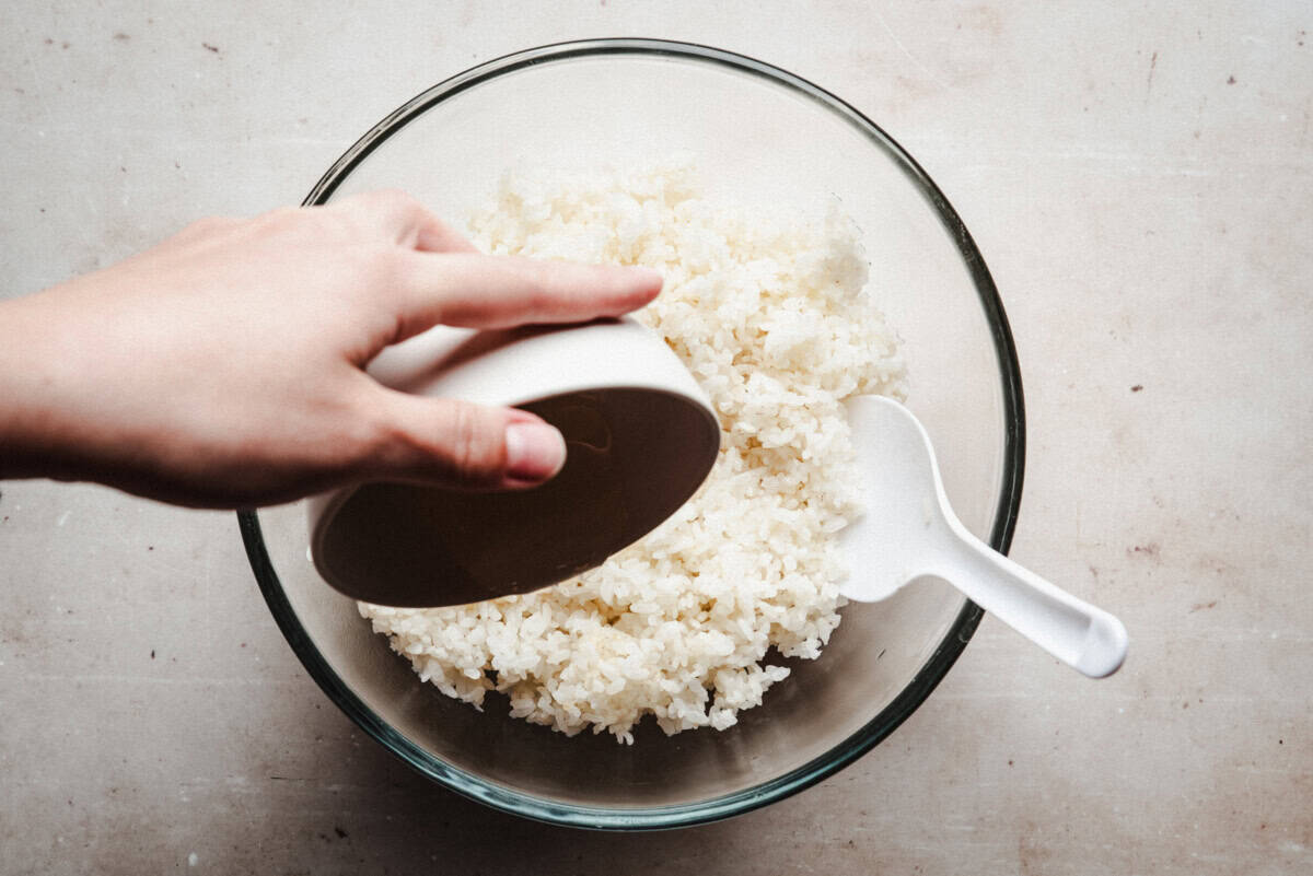 A hand pours liquid from a small bowl into a glass bowl of cooked short grain rice, with a white plastic rice paddle resting inside.