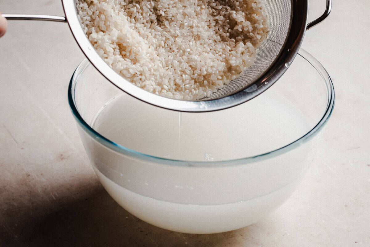 A metal strainer holding rinsed rice is positioned over a glass bowl filled with cloudy water, likely from washing the rice, on a light-colored surface.