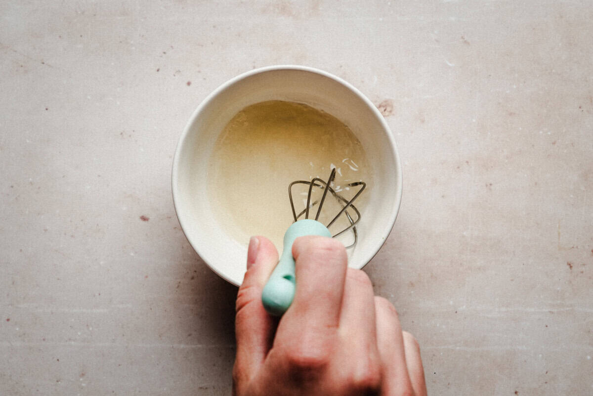 A hand holding a whisk with a light blue handle is mixing seasoning liquid in a white bowl on a light-colored surface.