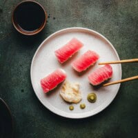 A plate with four pieces of tuna nigiri sushi, pickled ginger, and wasabi, with chopsticks picking up one piece. A small bowl of soy sauce is placed nearby on a dark green surface.