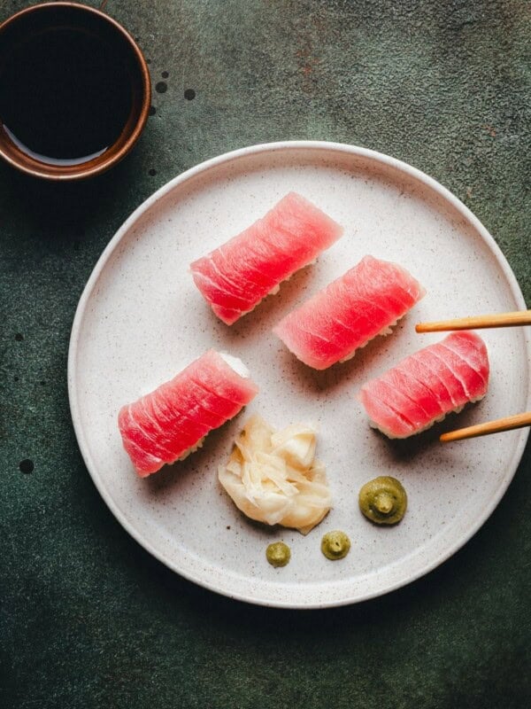 A plate with four pieces of tuna nigiri sushi, pickled ginger, and wasabi, with chopsticks picking up one piece. A small bowl of soy sauce is placed nearby on a dark green surface.