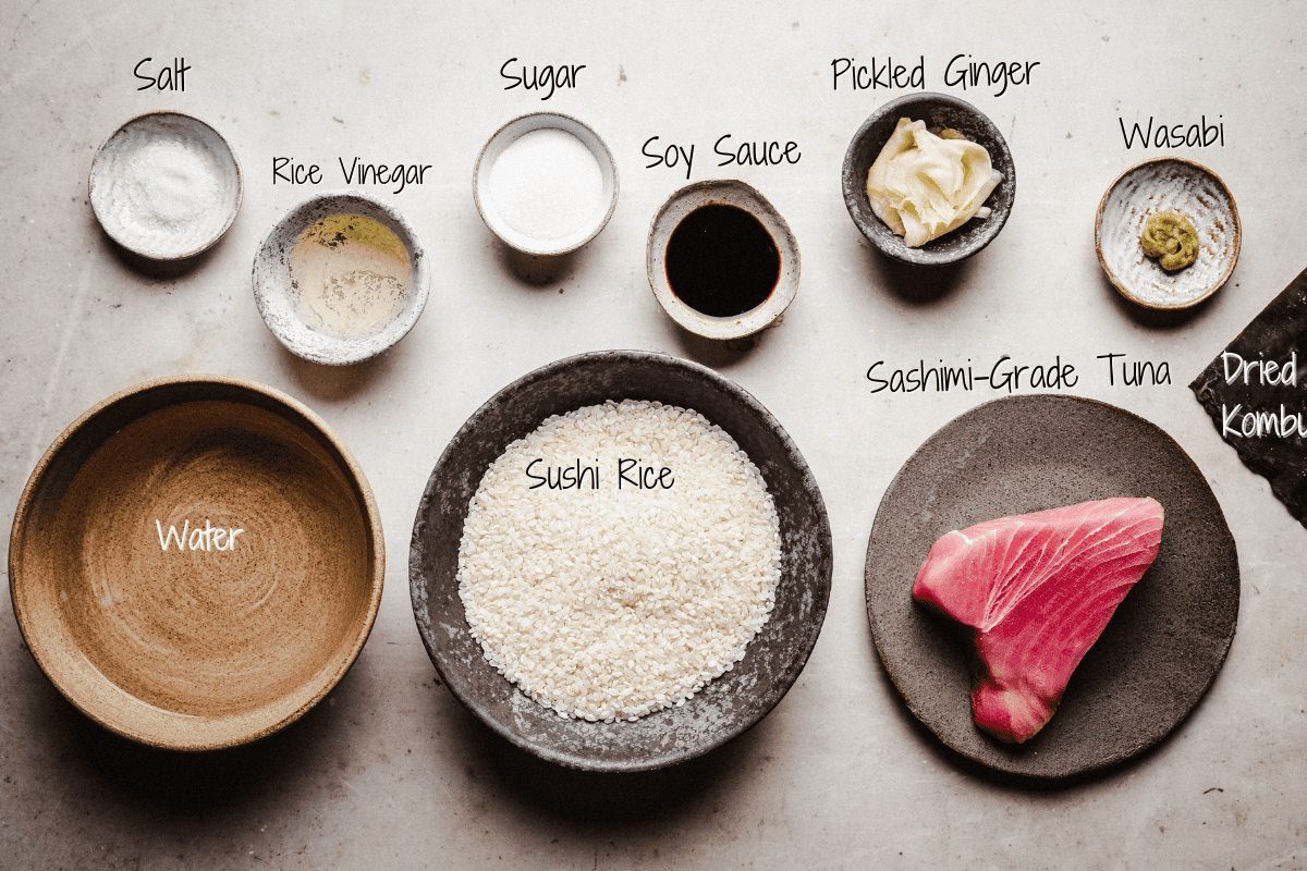 Top-down view of tuna nigiri ingredients, each labeled: salt, rice vinegar, sugar, soy sauce, pickled ginger, wasabi, dried kombu, water, sushi rice, and sashimi-grade tuna on various bowls and plates.