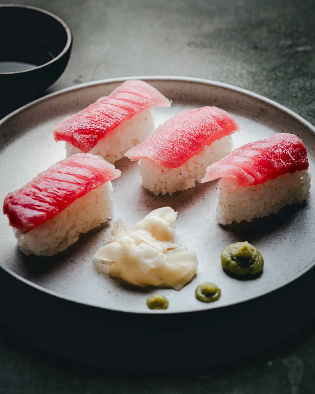 A plate with four pieces of tuna nigiri sushi, served with a small mound of pickled ginger and two dots of wasabi. A bowl of soy sauce is partially visible in the background.