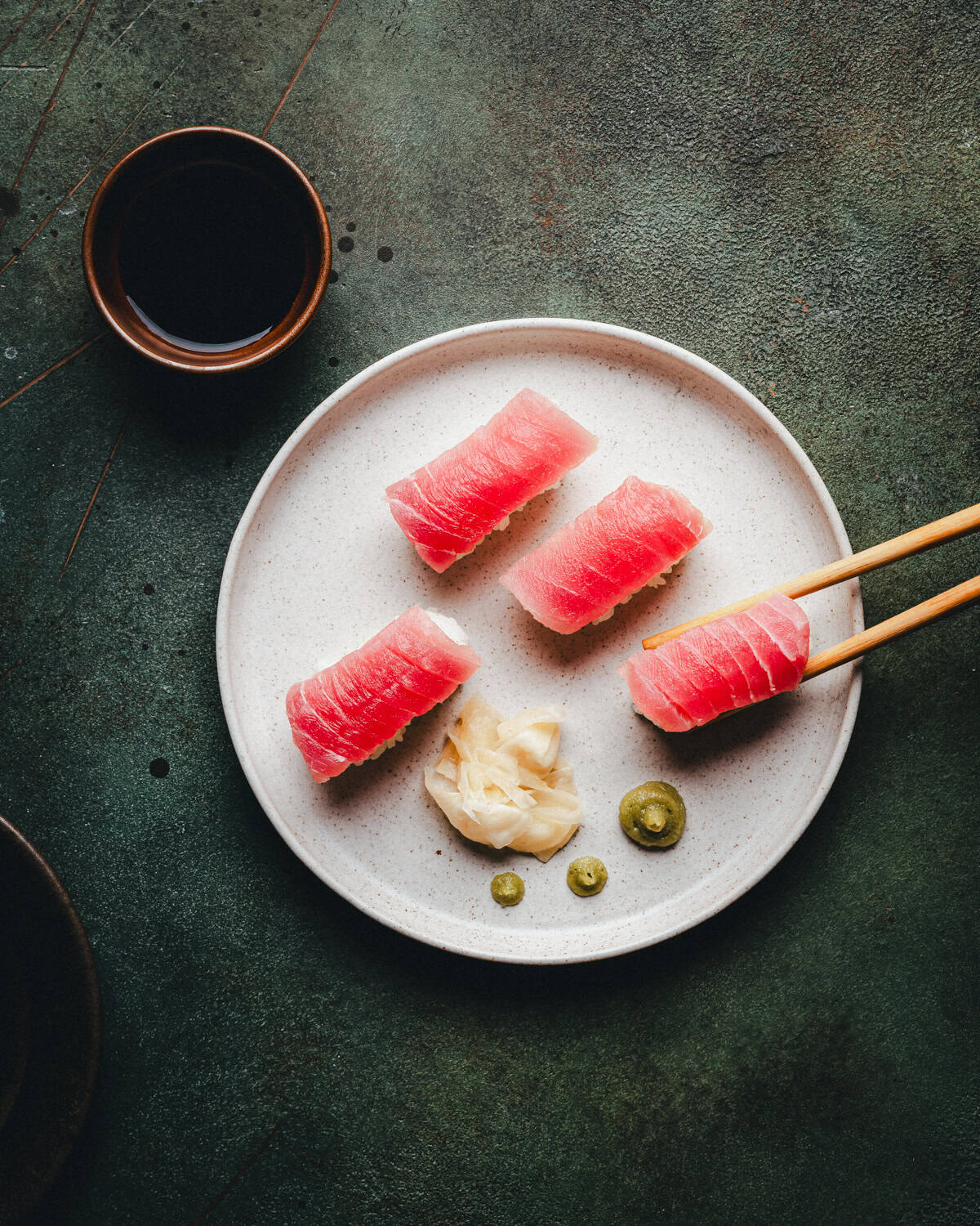 A white plate with four pieces of tuna nigiri, a serving of pickled ginger, and two small wasabi portions. Chopsticks hold one piece, and a cup of soy sauce sits nearby on a dark textured surface.