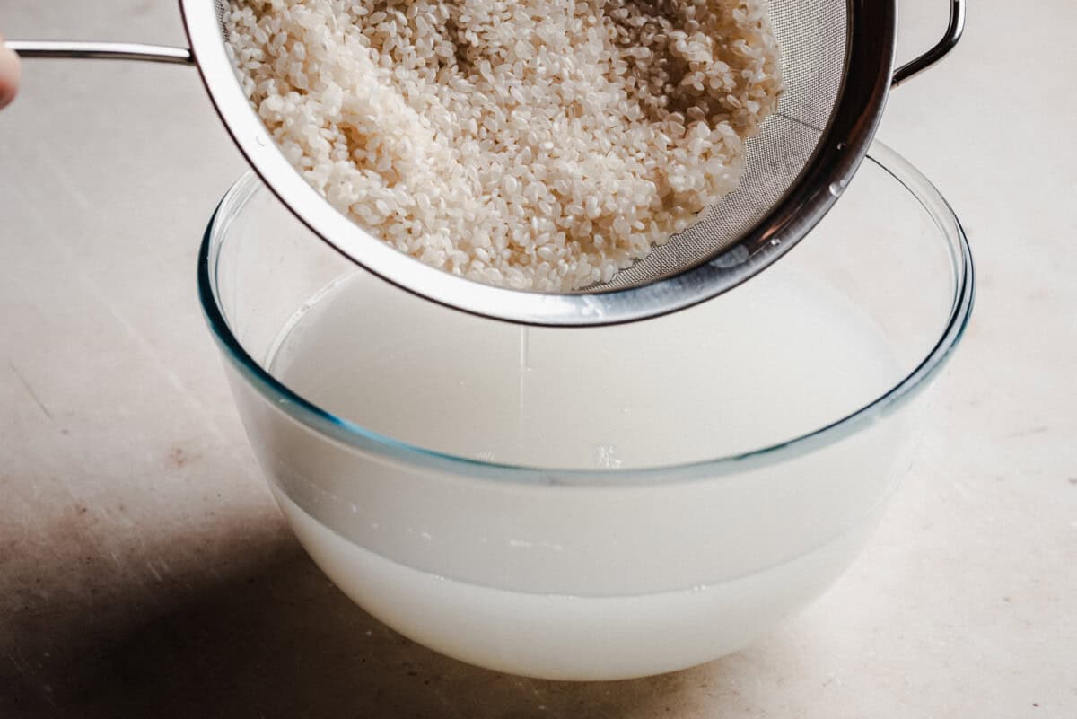 A metal strainer with rinsed rice is held above a glass bowl filled with cloudy water, showing the process of washing rice before cooking.