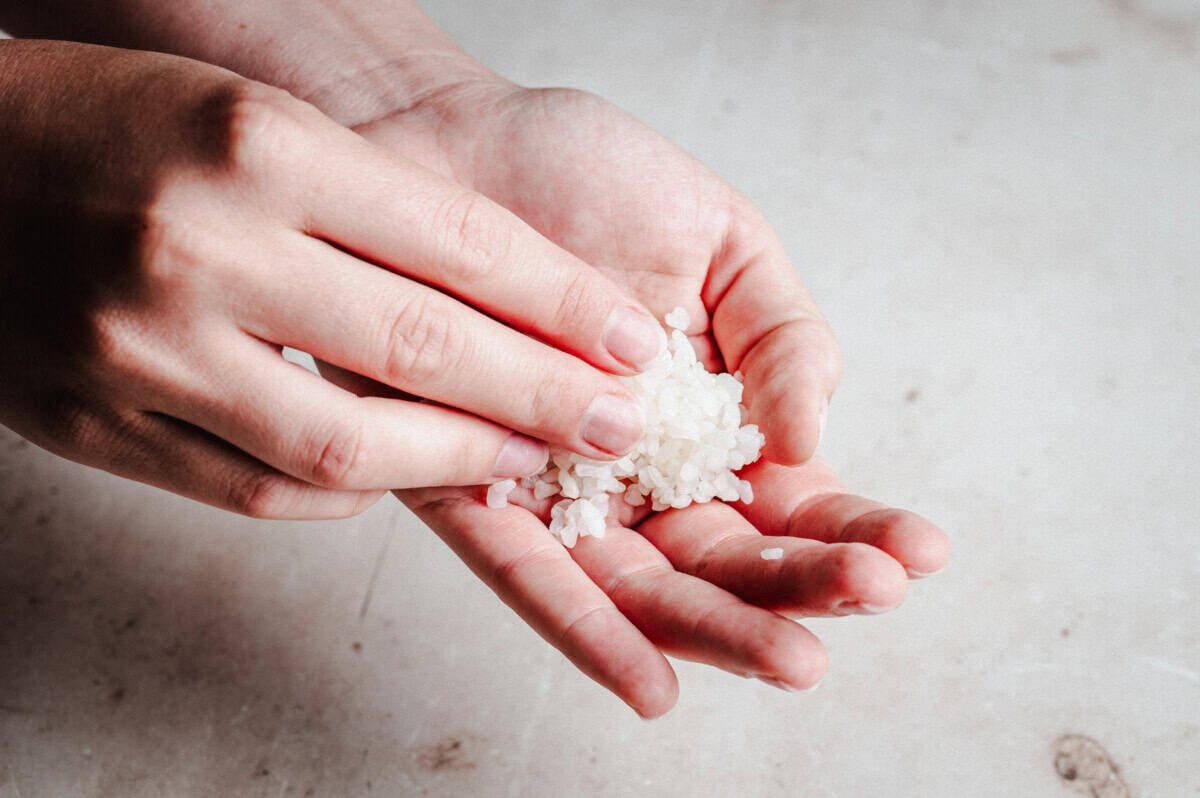 A person holding a small pile of uncooked white rice grains in one hand while gently touching the rice with the other hand, over a light-colored surface.