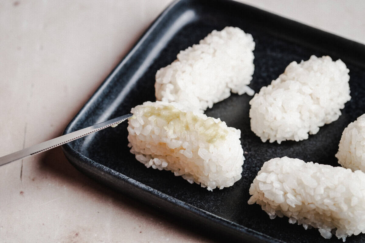 A close-up of rice balls arranged on a black square plate, with a small knife spreading wasabi on one of the rice balls.