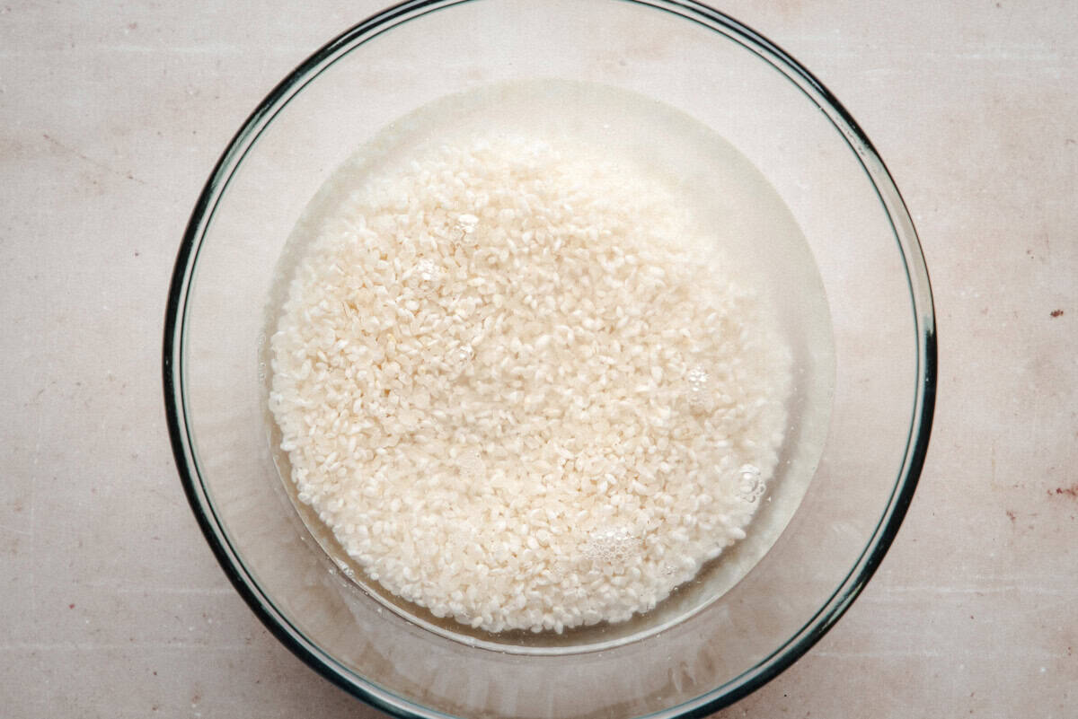 A glass bowl filled with uncooked white rice soaking in water, placed on a light-colored surface.