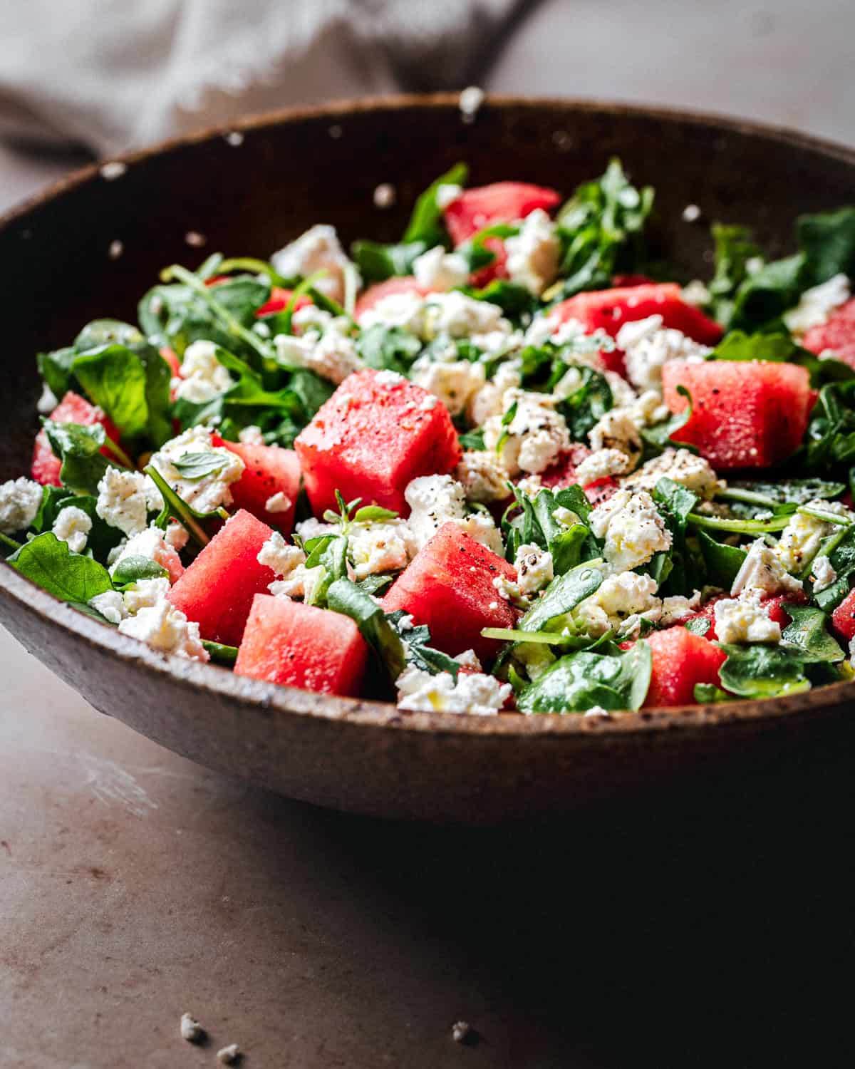 A close-up of a salad in a rustic brown bowl, featuring chunks of watermelon, crumbled feta cheese, and fresh green leaves, lightly seasoned with black pepper.