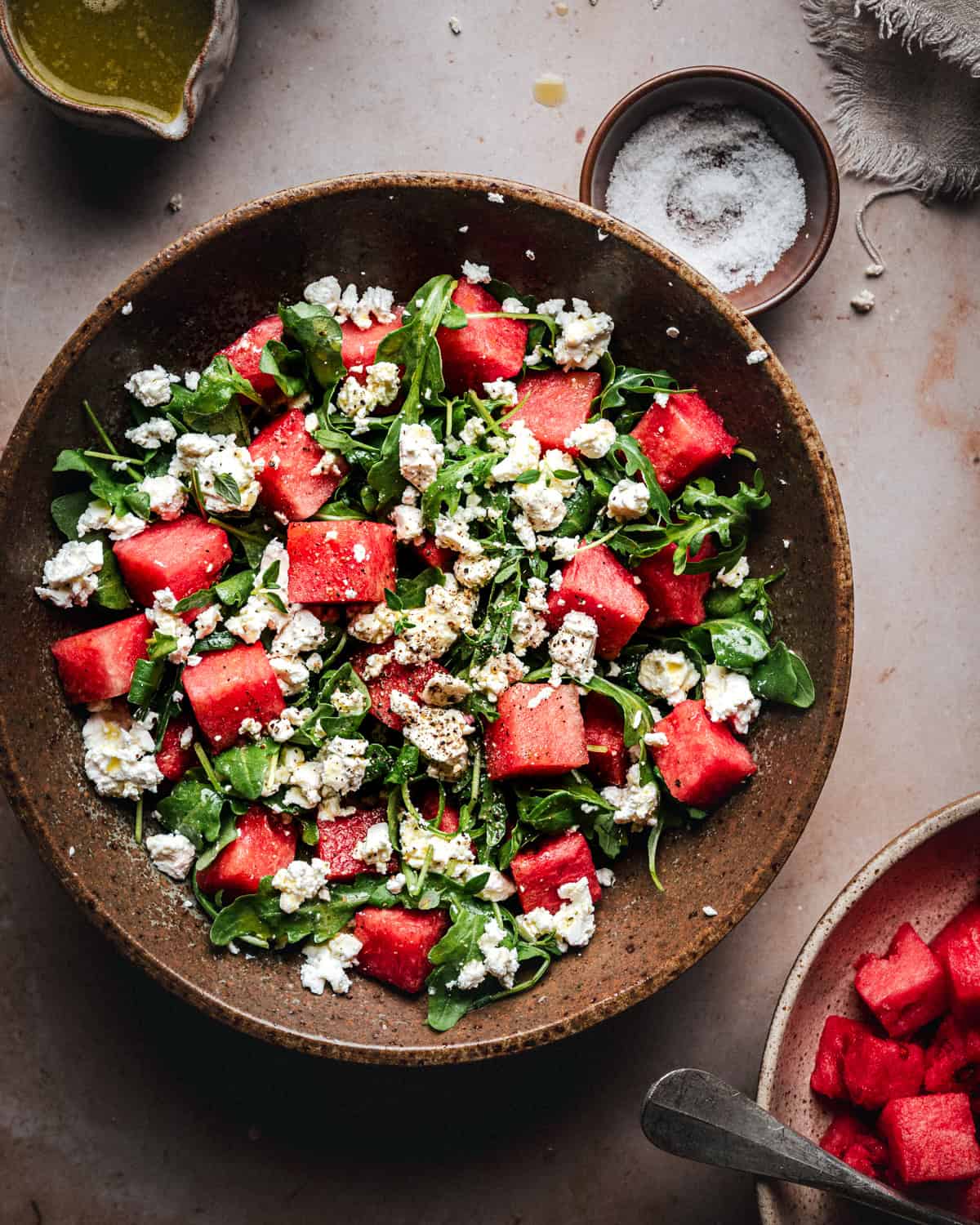 A brown bowl filled with a salad of watermelon cubes, arugula, and crumbled feta cheese, with a small bowl of salt, a bowl of olive oil, and extra watermelon pieces nearby on a light surface.
