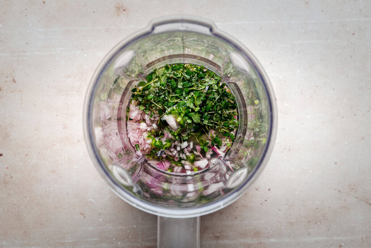 Chopped fresh herbs and diced onions inside a blender viewed from above, ready to be blended on a light-colored countertop.