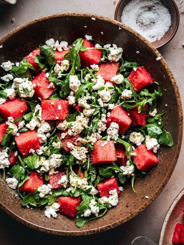 A bowl of fresh salad with chunks of watermelon, arugula leaves, and crumbled feta cheese, topped with ground black pepper, next to a small bowl of salt.
