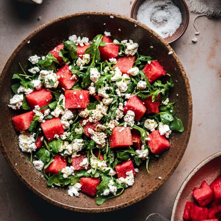 A bowl of fresh salad with chunks of watermelon, arugula leaves, and crumbled feta cheese, topped with ground black pepper, next to a small bowl of salt.
