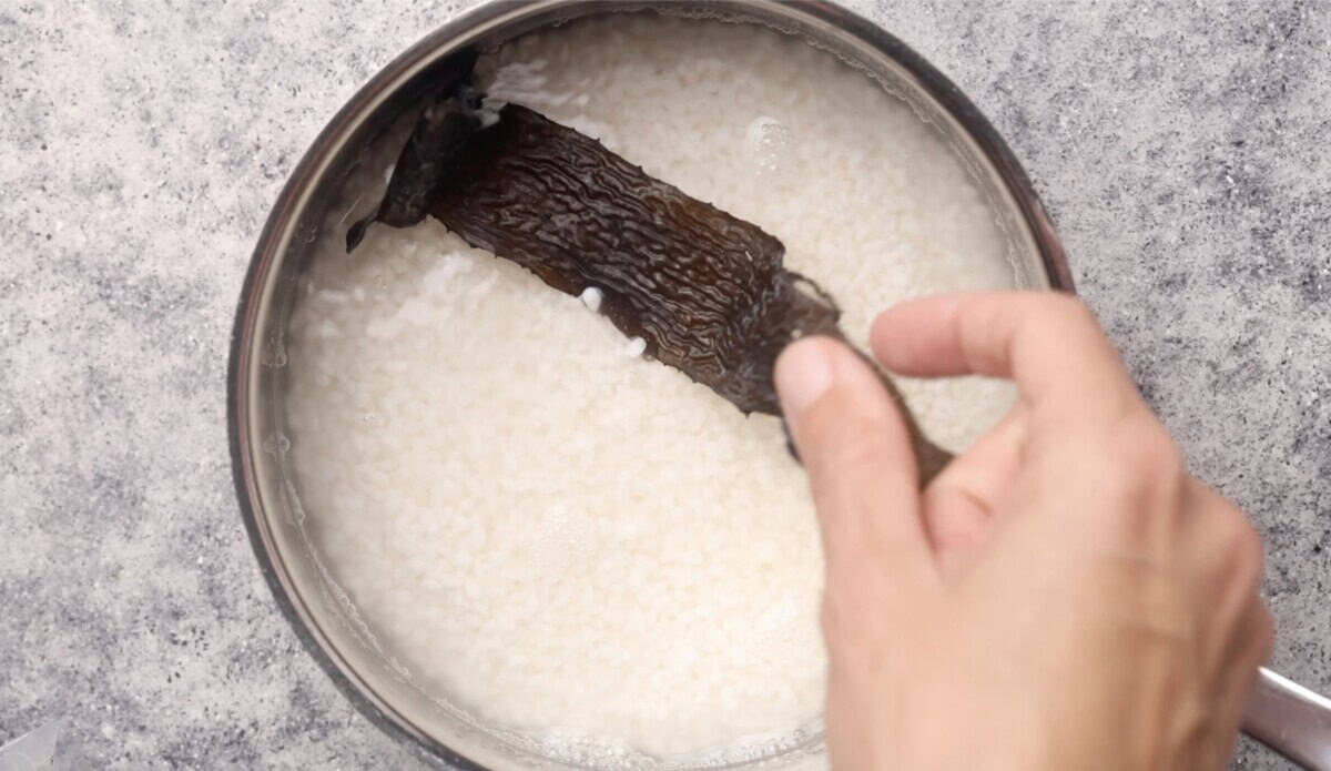 A hand places a piece of dried kombu seaweed into a pot of soaking white rice in water, preparing to make sushi rice on the stovetop.