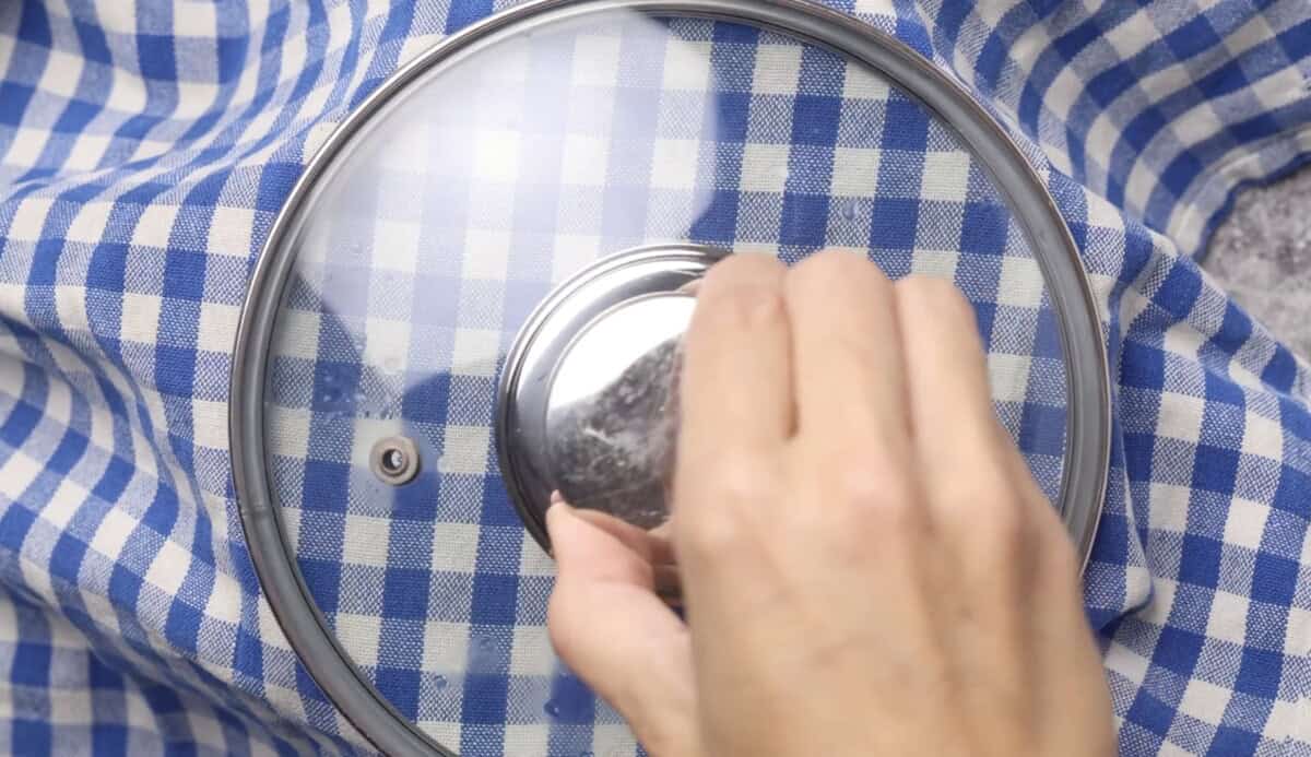 A hand lifts the glass lid of a pot, which is placed on a blue and white checkered cloth.