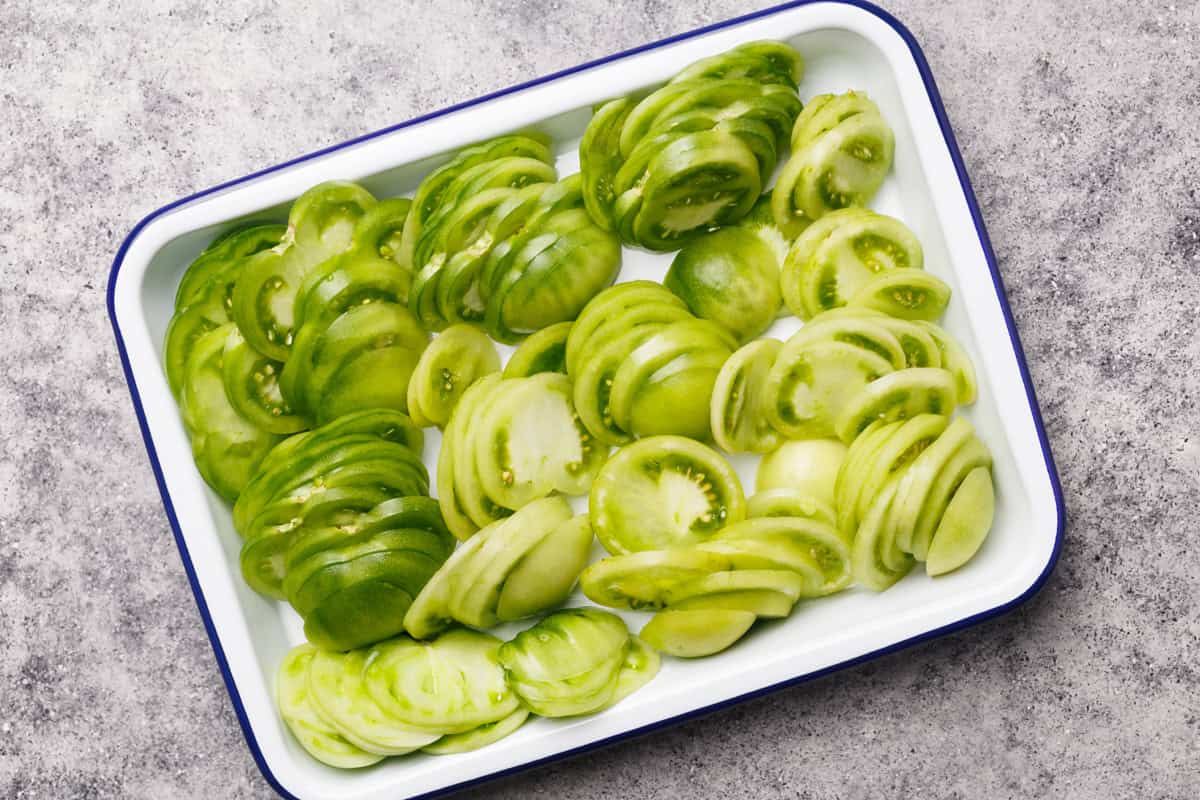 A white enamel tray with a blue rim holds several rows of thinly sliced green tomatoes, arranged in an overlapping pattern on a gray stone surface.