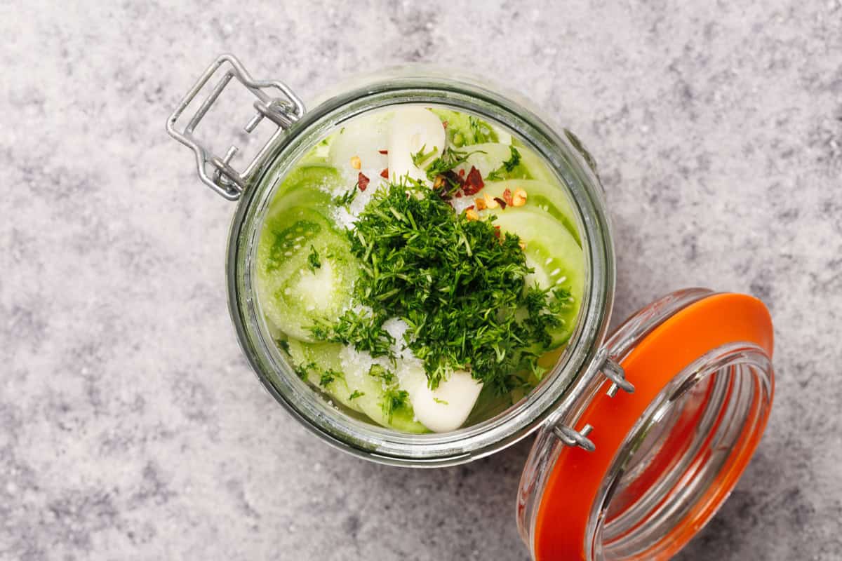 Open glass jar filled with sliced green tomatoes, garlic, fresh herbs, red pepper flakes, and salt, ready for pickling, on a gray textured surface. The jar lid is open and rests to the side.