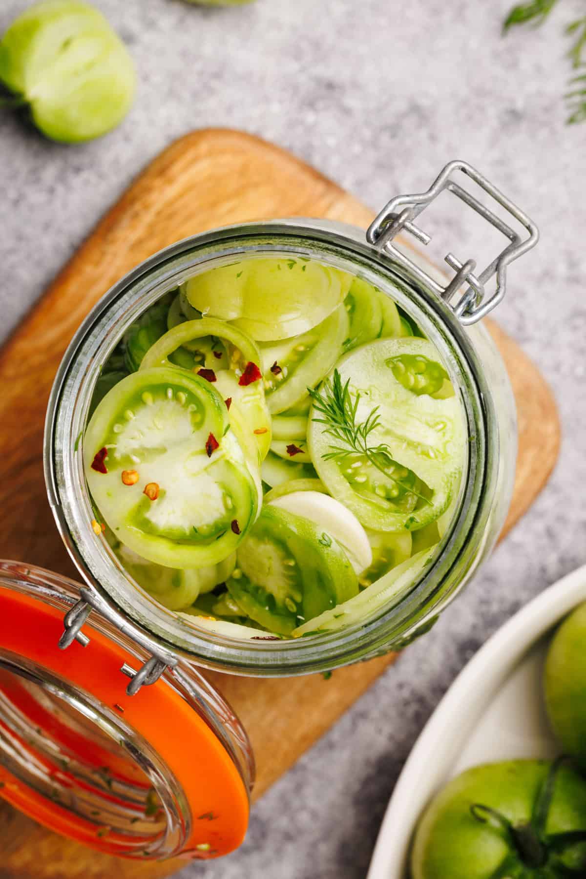 Open glass jar filled with sliced green tomatoes, onions, dill, and red pepper flakes, sitting on a wooden cutting board with whole green tomatoes nearby.