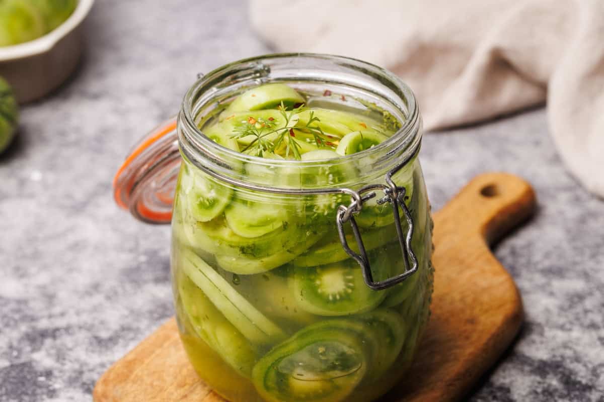 A glass jar filled with sliced green tomatoes, herbs, and pickling liquid sits on a wooden cutting board, with a gray cloth in the background.