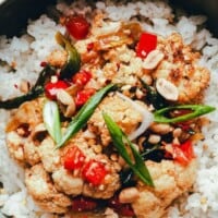 A bowl of white rice topped with kung pao cauliflower, featuring red bell peppers, green onions, peanuts, and a savory sauce. Text reads: โKung Pao Cauliflower - pinchandswirl.com.โ.