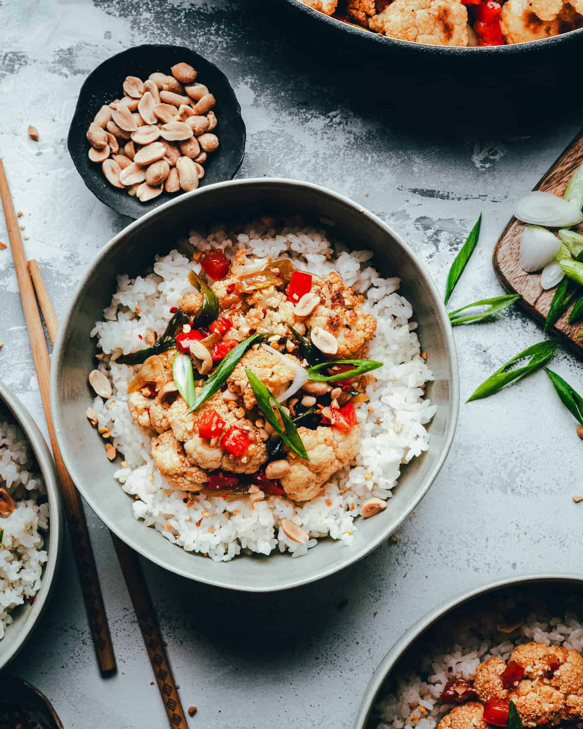 A bowl of white rice topped with kung pao cauliflower and garnished with chopped peanuts and green onions, with chopsticks, a dish of peanuts, and more ingredients on the table nearby.