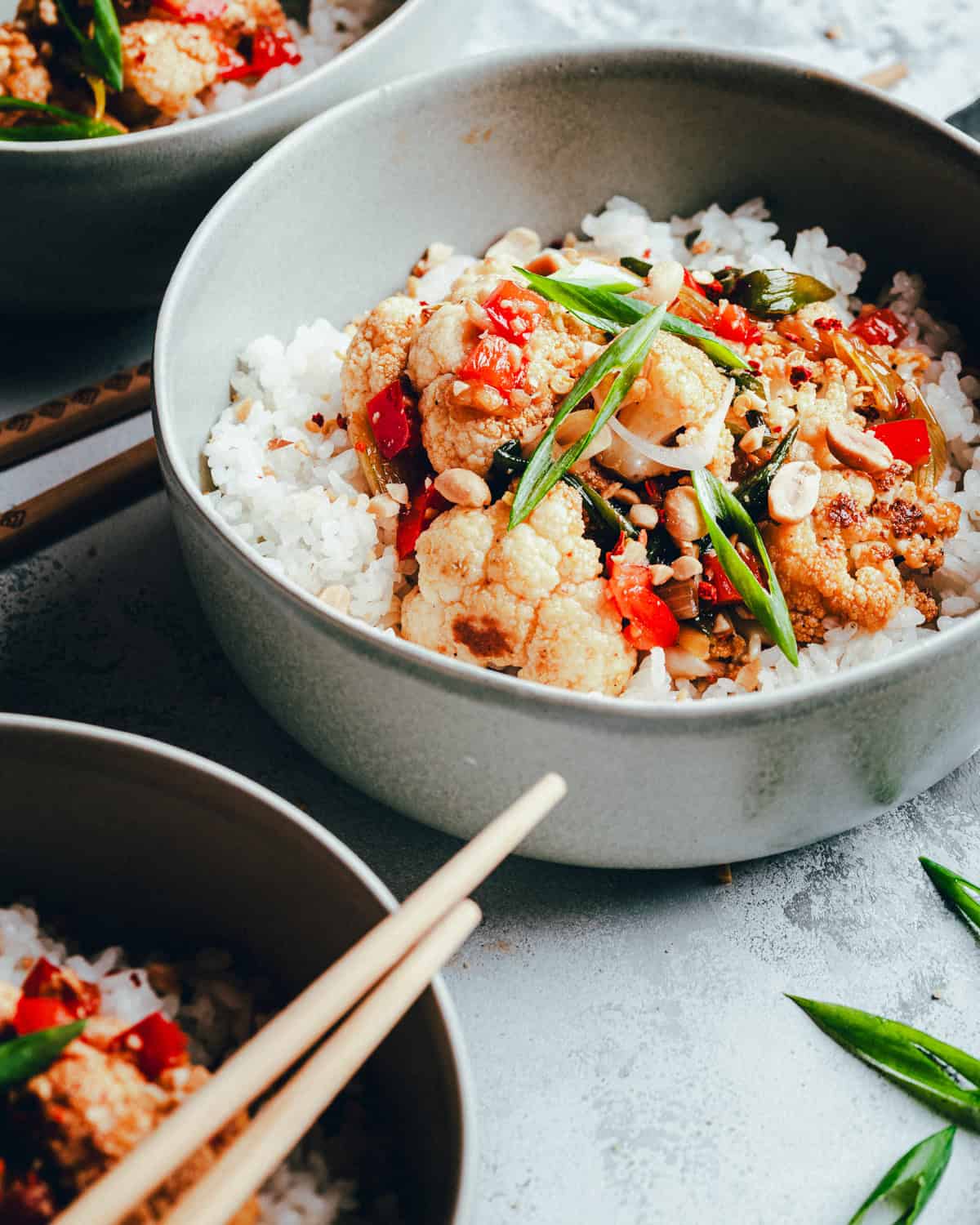 A bowl of white rice topped with kung pao cauliflower, red peppers, green onions, and chopped peanuts, with chopsticks resting on the side.