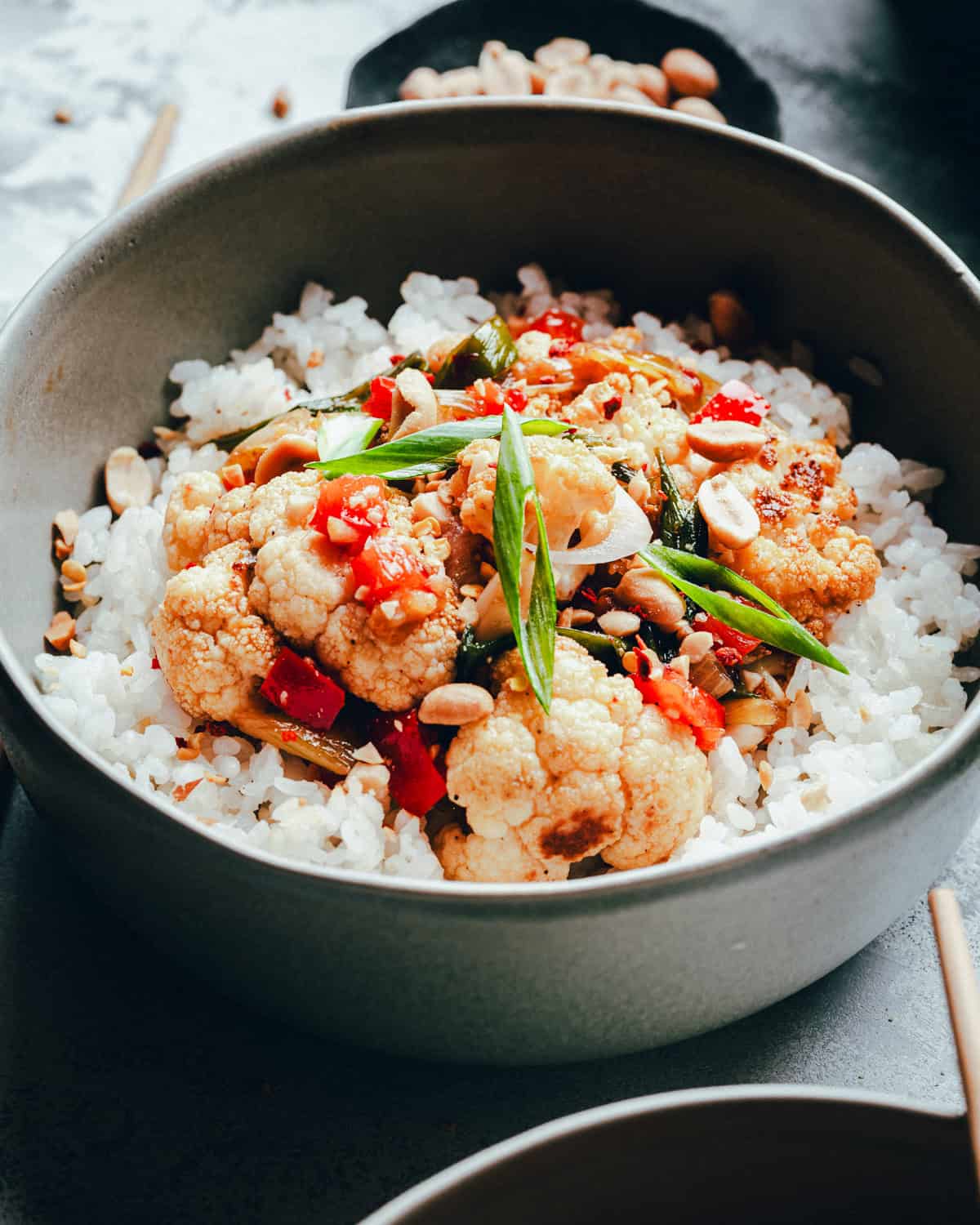 A bowl of white rice topped with kung pao cauliflower, chopped red peppers, green onions, and peanuts, served in a dark bowl with chopsticks nearby.