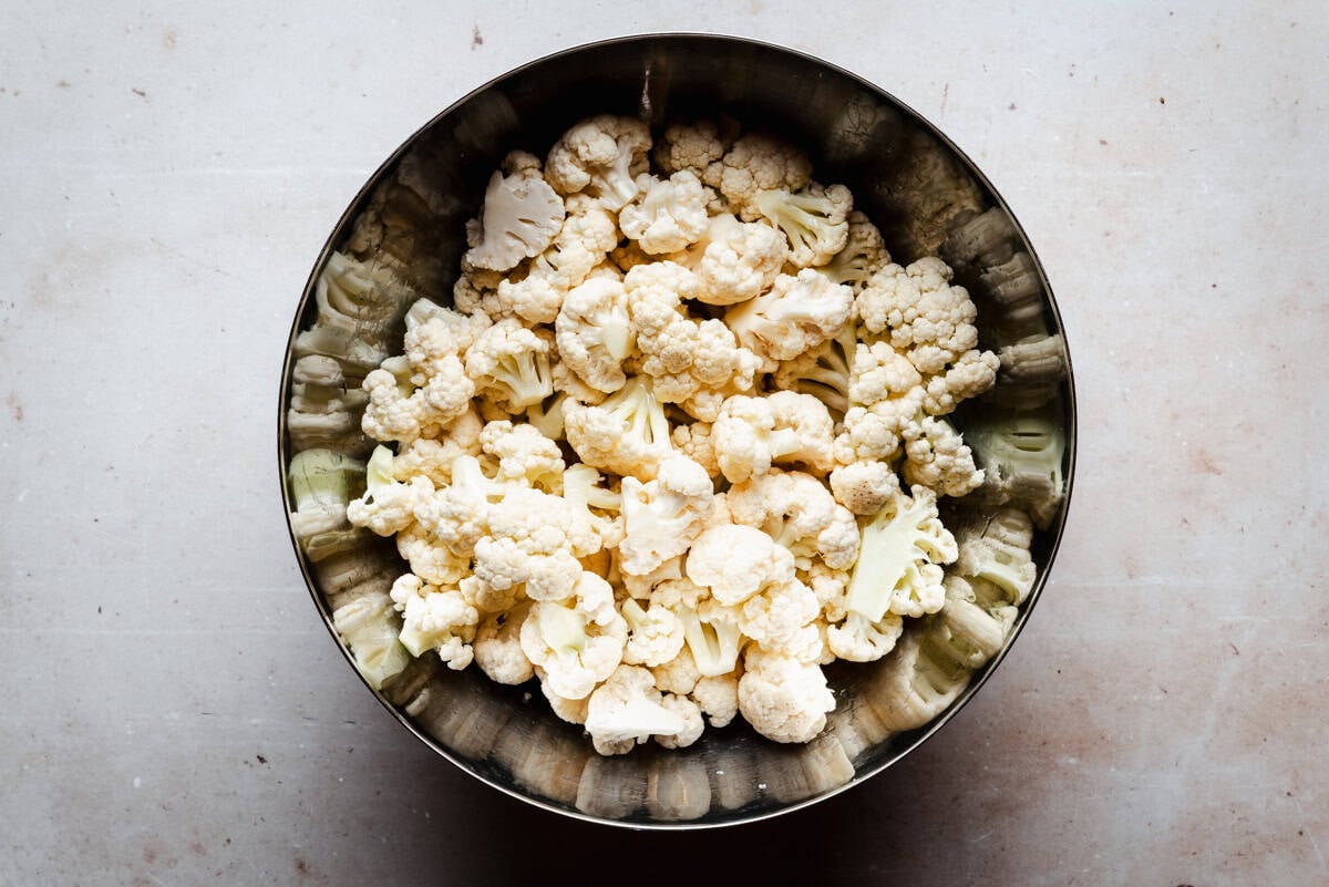 A metal bowl filled with fresh cauliflower florets sits on a light, speckled countertop, viewed from above.