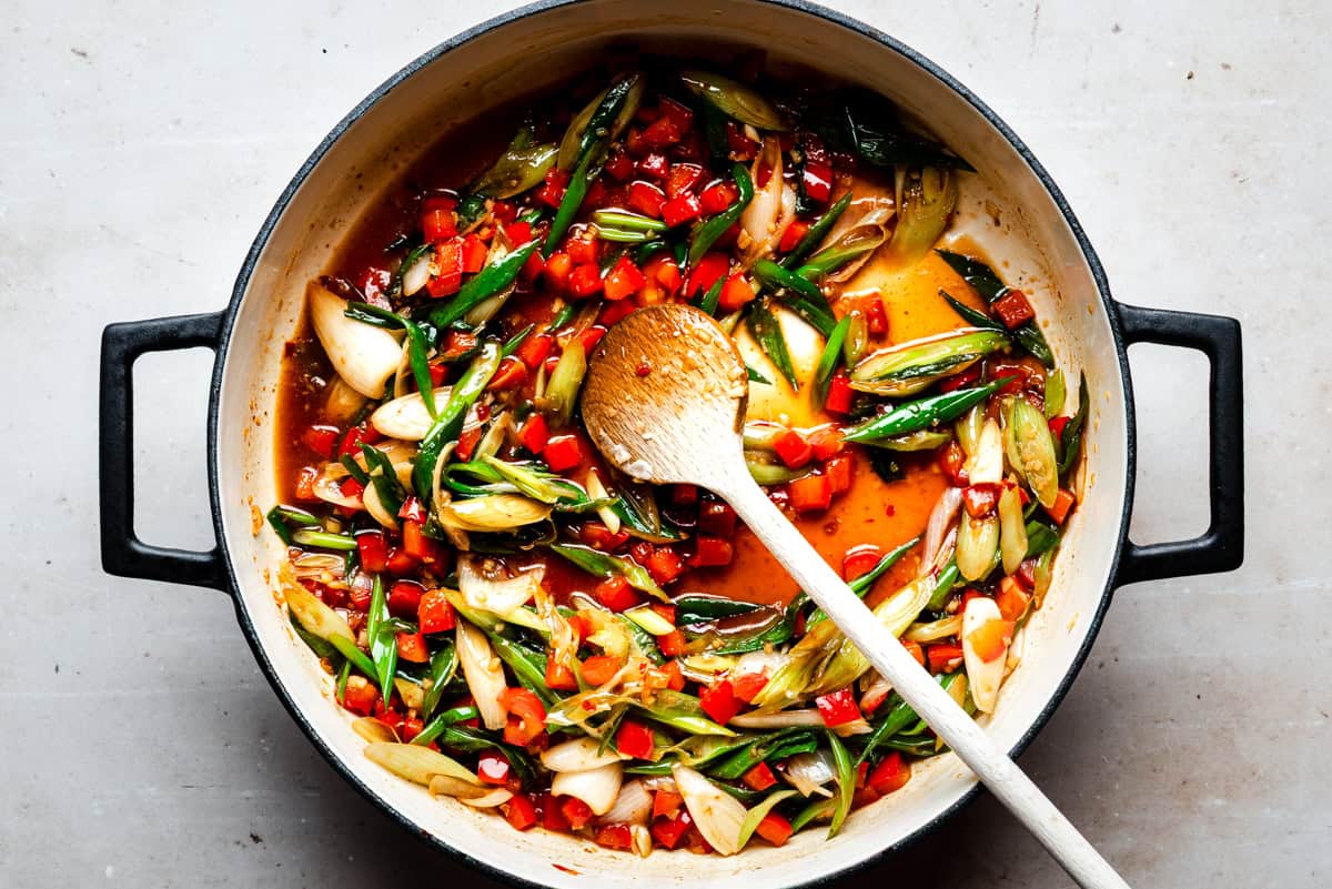 A pot filled with a colorful mix of sautéed vegetables, including sliced green onions, red bell peppers, and a rich, orange-red sauce. A wooden spoon rests in the pot, stirring the ingredients.