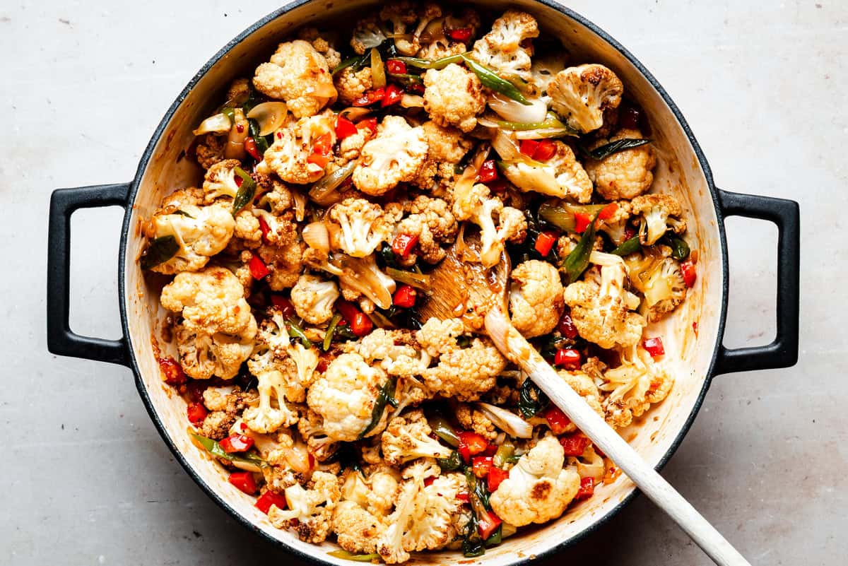 A large pot filled with cooked cauliflower florets, red bell peppers, onions, and green herbs, all mixed together with spices. A wooden spoon rests inside the pot on a light-colored surface.